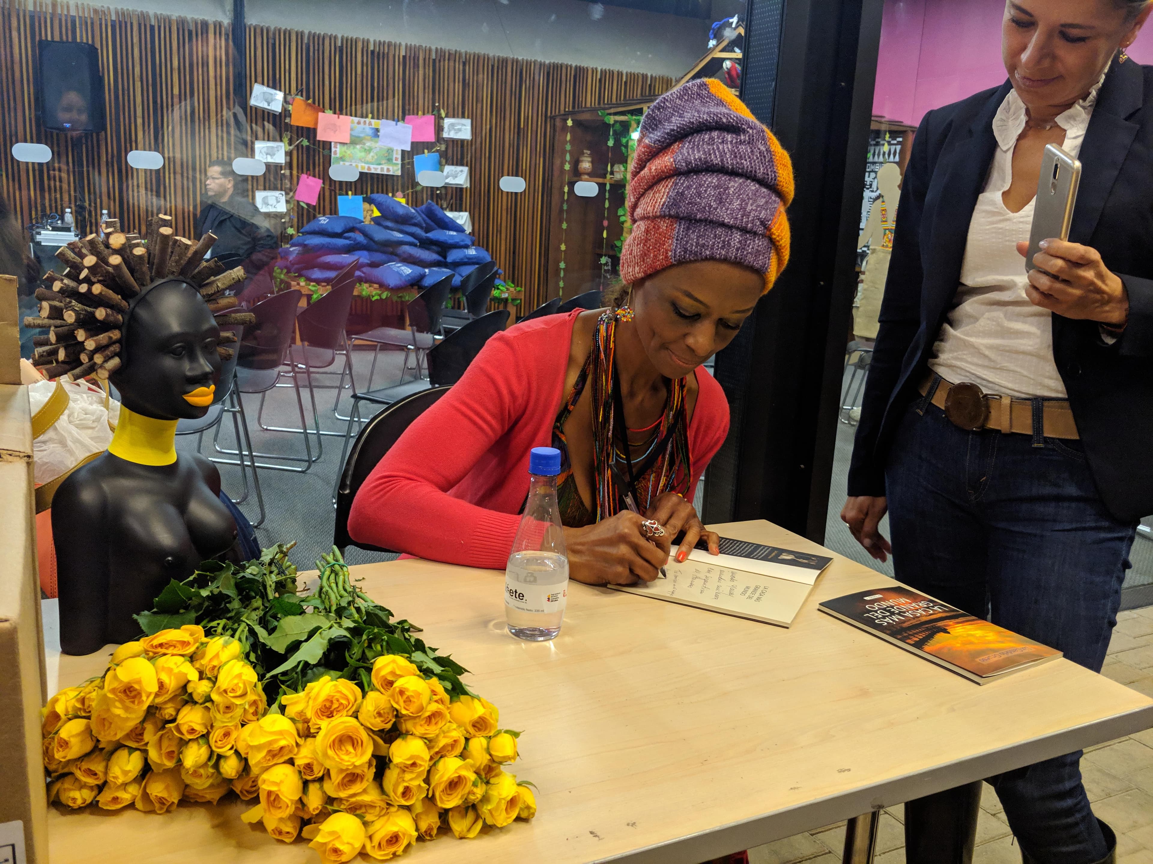 Liz Candelo firmando un libro en la FILBo 2019, con un busto decorativo y rosas amarillas sobre la mesa